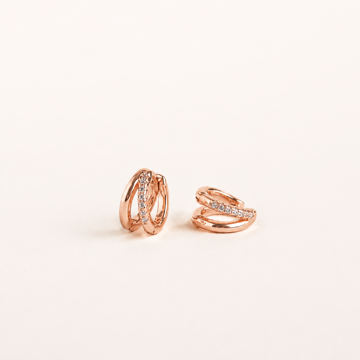 A pair of rose gold huggie hoop earrings with small clear pavé-set stones, displayed on a clean off-white background.